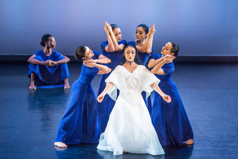 Four women in blue dress crowd around a woman in white. Their arms touch in front of their face with their hands in a prayer position. A sits in the background.